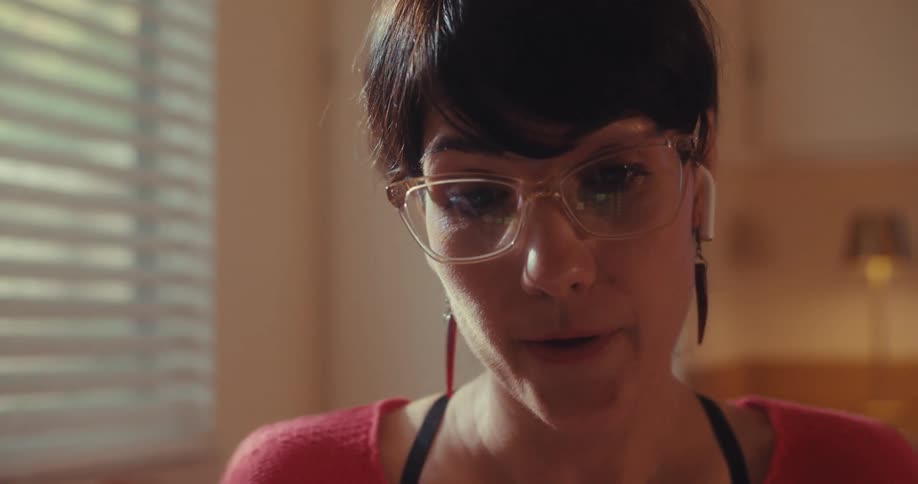 The face of a woman who is wearing reading glasses and wireless headphones and she is talking looking at something that is below her
