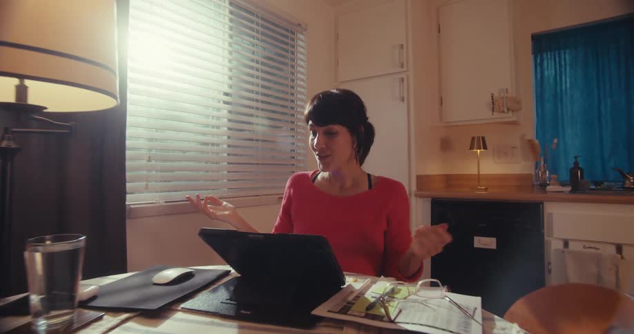 A woman sits at a kitchen table on a video call using a laptop