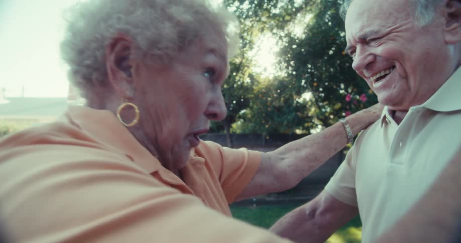 An elderly couple smiles while dancing together in a sunny backyard