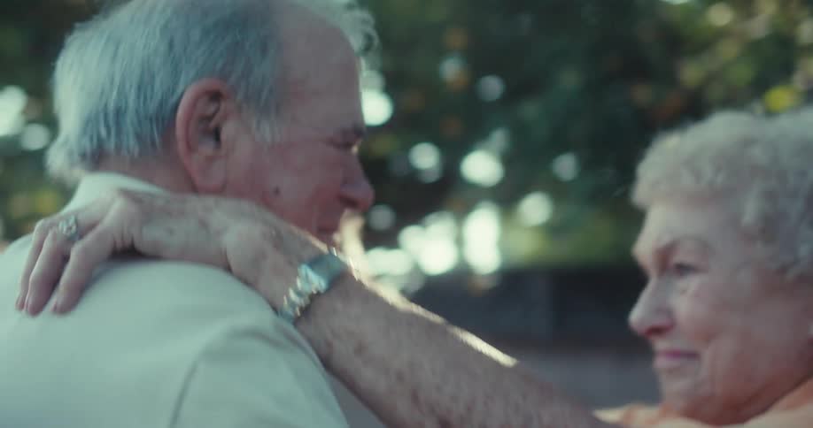 An elderly couple dancing together near a flowering tree under sunlight