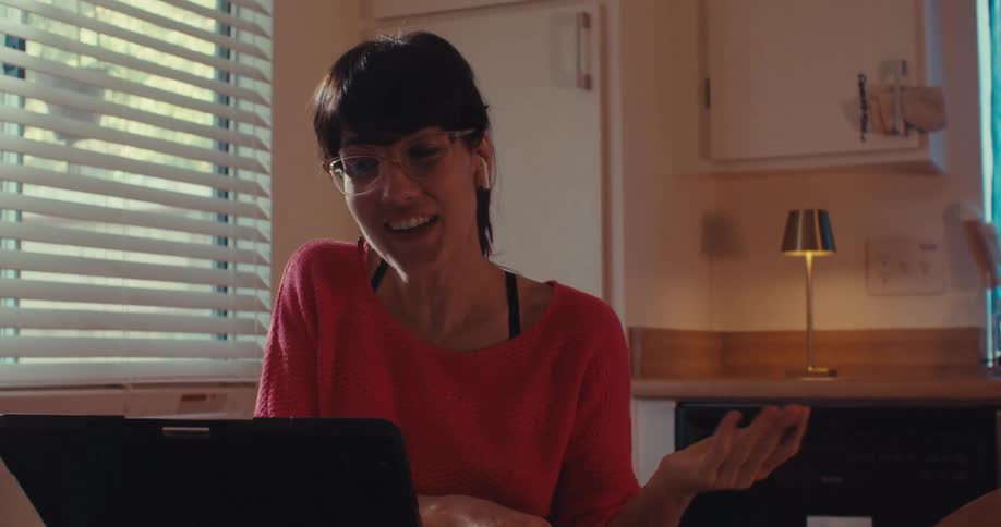 A woman in a red sweater sits at a kitchen table writing on paper with a laptop open and a lamp lit behind her
