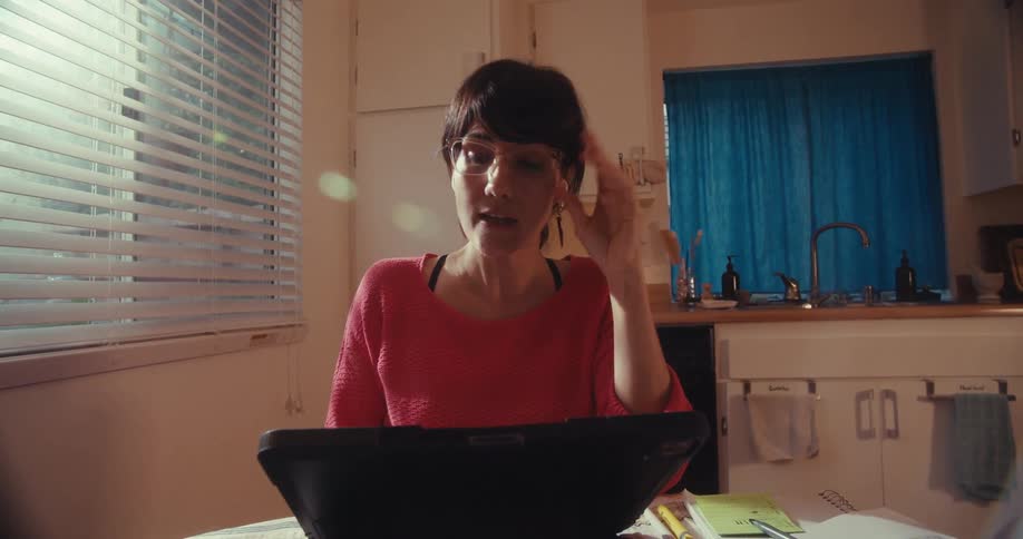 Woman in glasses sits at a kitchen table on a video call discussing workrelated stress