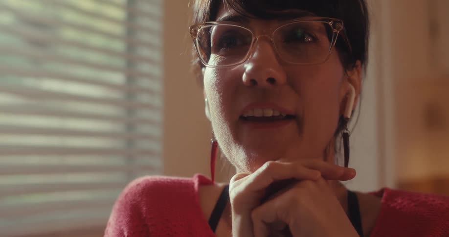 A young woman wearing earbuds and glasses sits beside a window around midday lost in thought then laughs afterward
