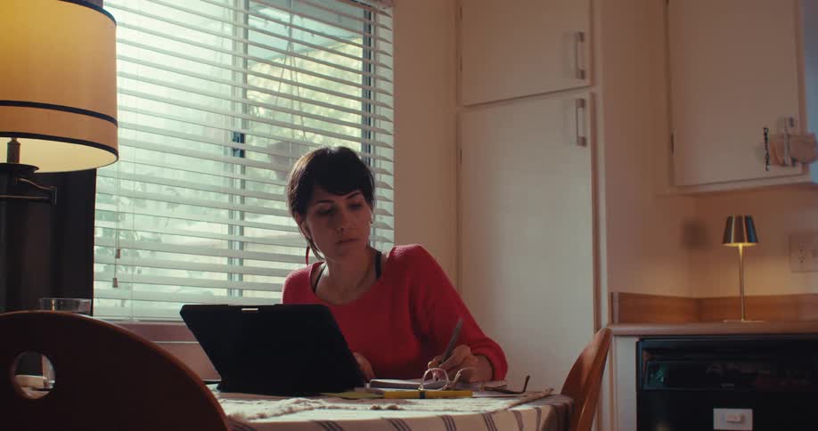 A woman is working at a kitchen table with a laptop and notepad