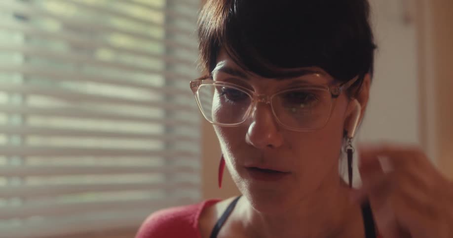 A woman wearing earbuds and glasses is on a call sitting beside a window with blinds around midday