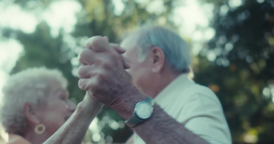 An elderly couple dances joyfully outdoors in warm sunlight