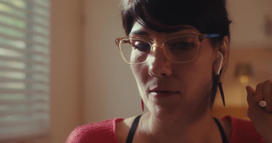 A woman with glasses and an earbud talks engagingly during a video call her face illuminated by soft light from a nearby window