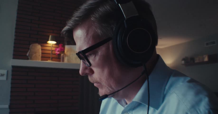 A man wearing headphones is sitting in a chair typing on a laptop in front of him in a living room