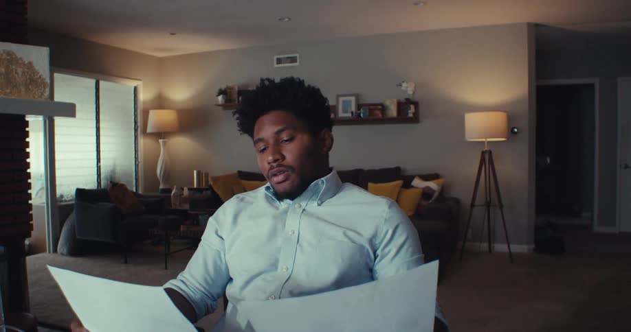 A man in a living room looks very stressed while shuffling papers drinking and talking on the phone