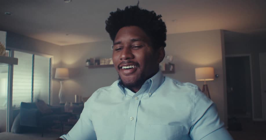 A man in a light blue shirt sits in a warmly lit room smiling and laughing