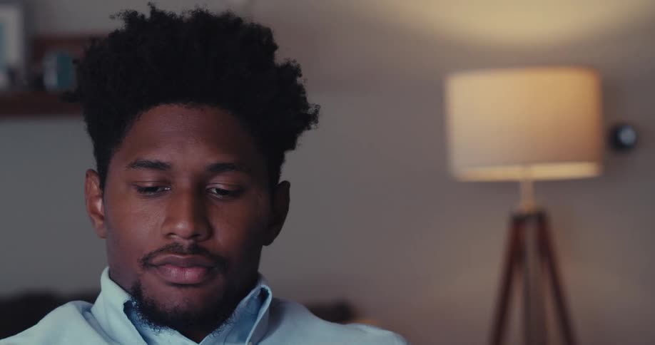 A young black man sits indoors in deep thought with a lamp behind him