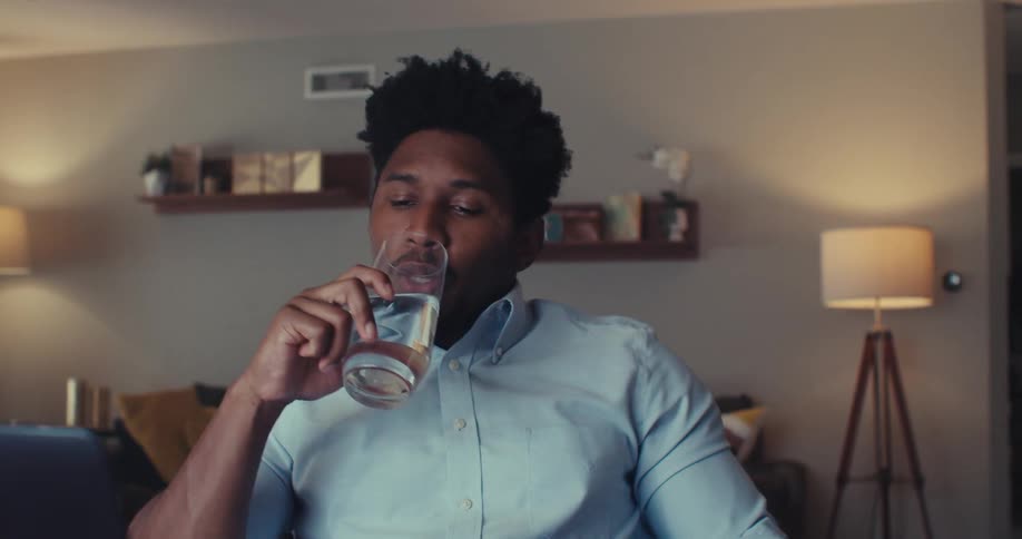 A young black man sits in front of a computer drinks a glass of water then continues his video conversation