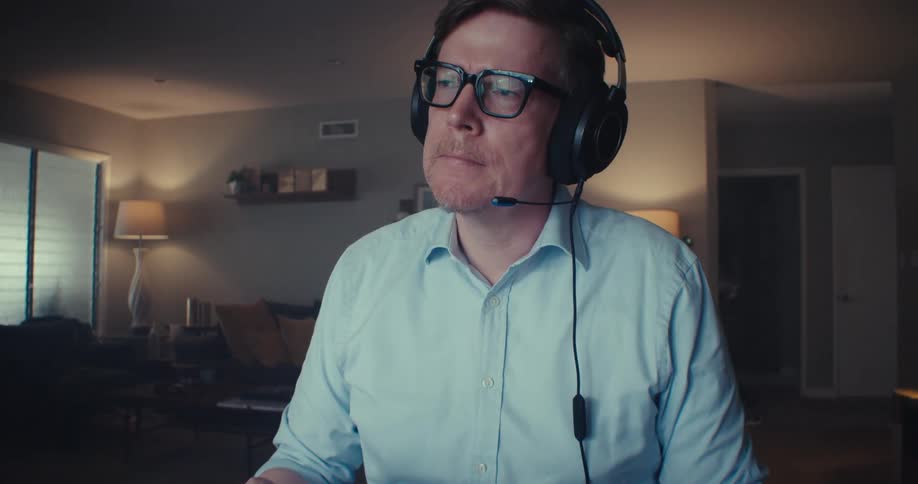 A man wearing headphones and is sitting in front of a table with laptop and he looks to the side in thoughtful tones