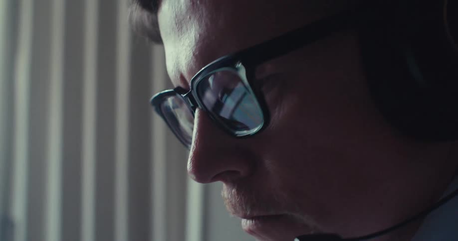 The face of a man wearing reading glasses and a headphone on his head and he gives a subtle smile while looking to the side