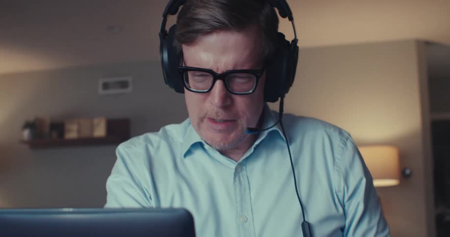 A man wearing headphones sits at a desk at home analyzing documents in front of a laptop