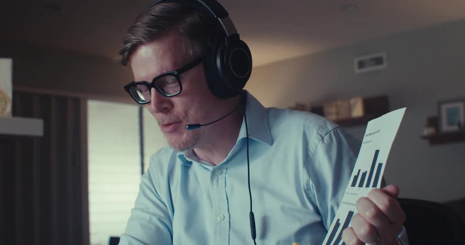 A man working from home wearing headphones holds a document with bar graphs while on a call at a desk with a laptop and yellow pen