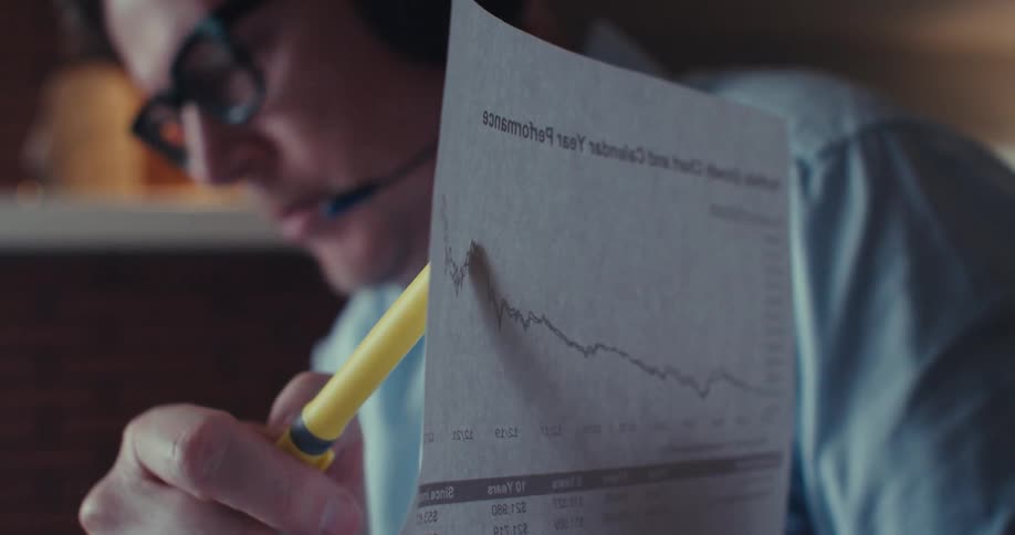 A man wearing headphones is pointing a yellow pen at a paper with graphs while talking
