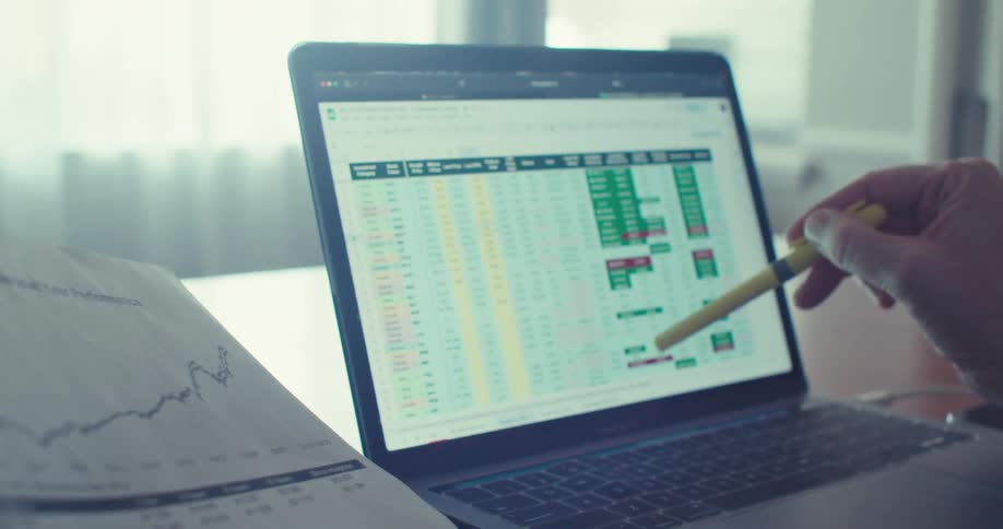 A hand holding a yellow highlighter hovers over a detailed spreadsheet on a laptop screen filled with colorcoded data points