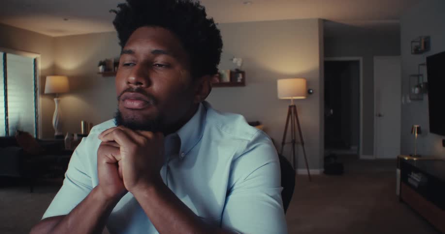 A young black man sits in his living room with both hands under his chin looking sad