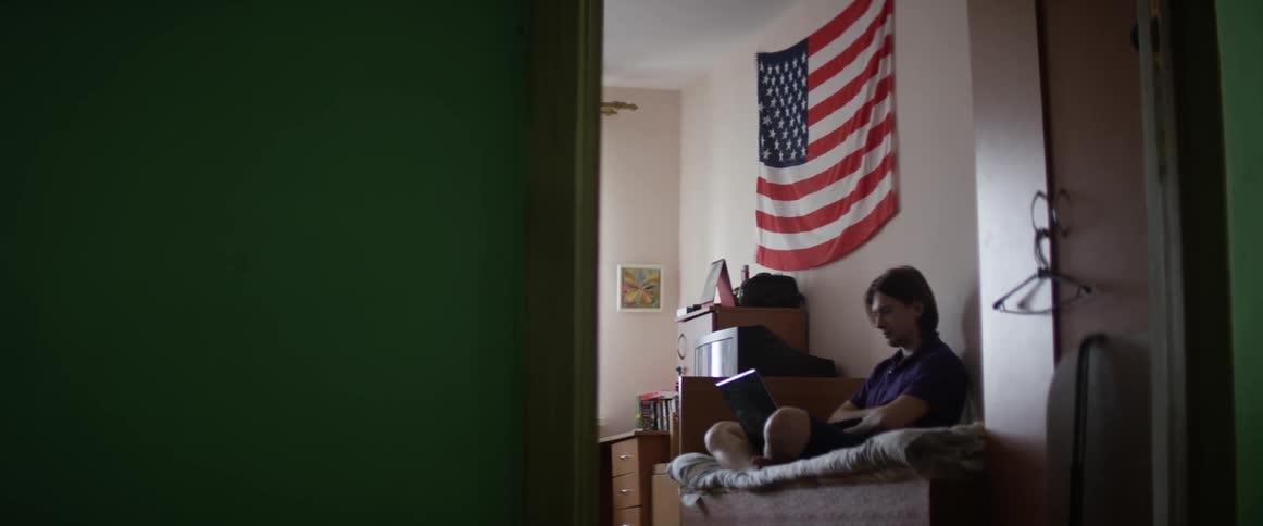 A man sits on a bed in a room watching something on a laptop