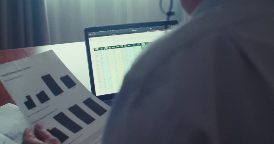 A man holds a document while wearing headphones with a microphone speaking into the mic as he looks at stock prices on a laptop monitor around midday at an office desk