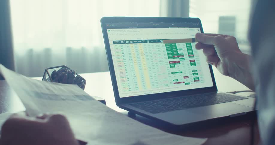 A man wearing a headset examines stock prices comparing them with a document he is holding around midday in his office