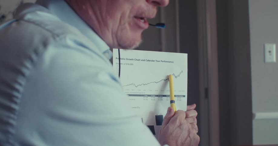 A man is using a pen to point to a paper with a graph and he is looking at a laptop screen
