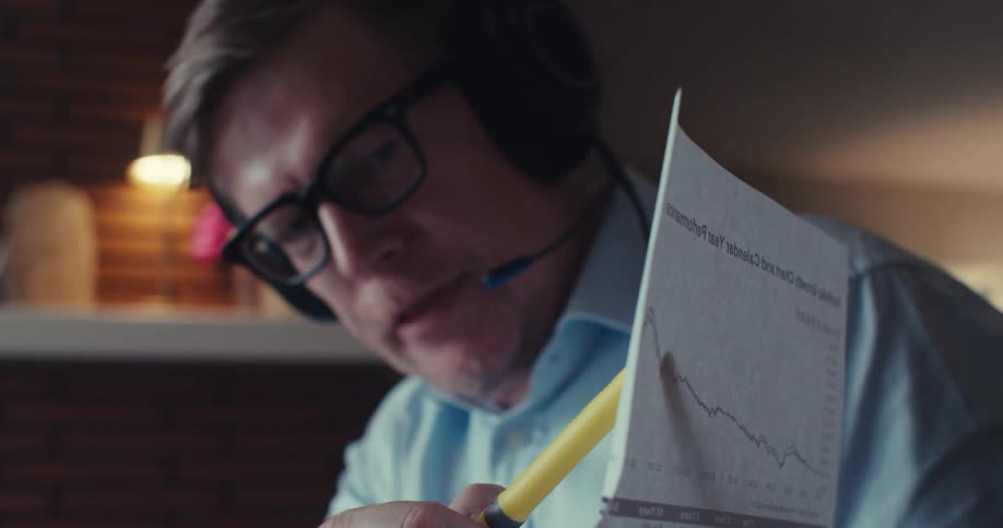 A man wearing a headset and glasses discusses a downward trending financial chart hes holding up during a video call