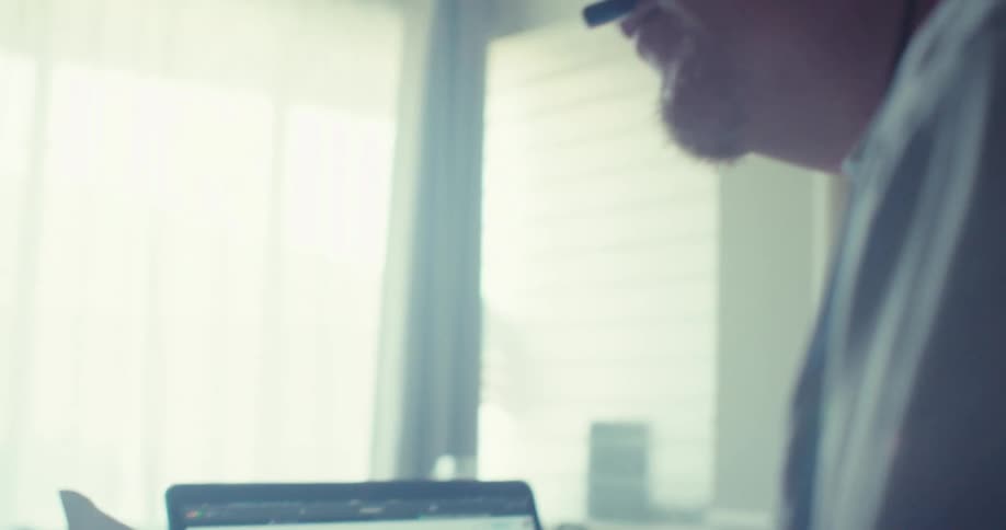 Close up of a man wearing headphones reading a document beside a laptop on a desk