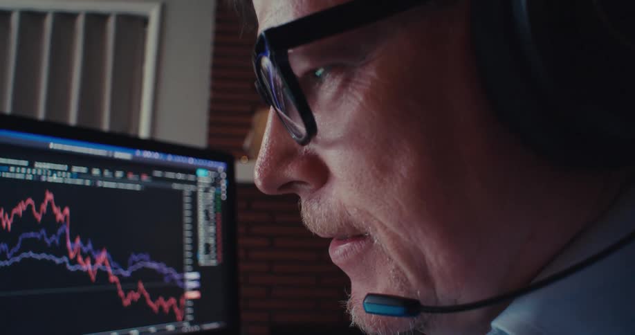 A man wearing headphones focuses on a computer screen showing stock market graphs