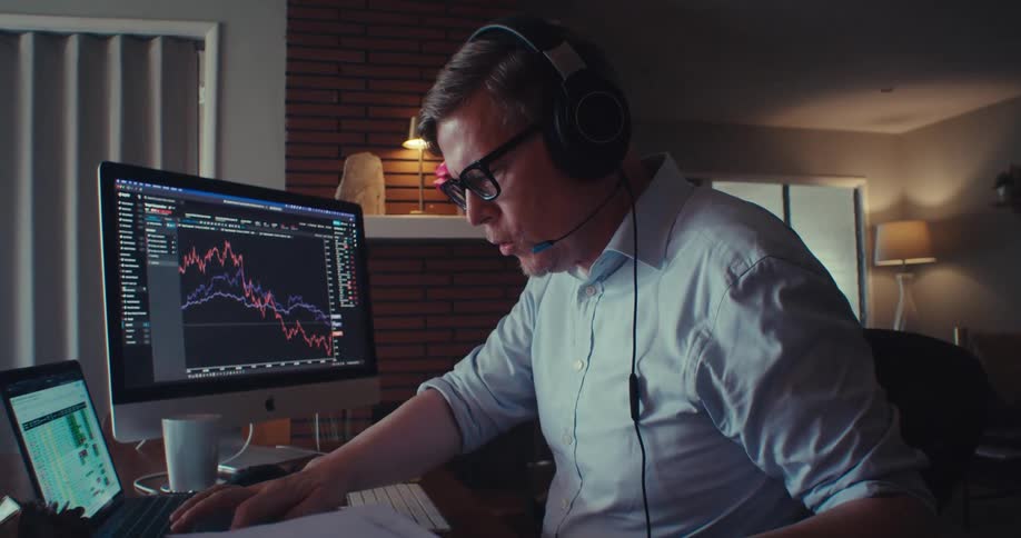 A man wearing a headset works from his home office at night analyzing stock market charts on his computer
