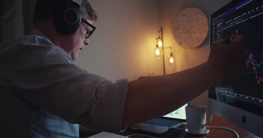 A man wearing headphones sits at a desk analyzing stock market graphs on a computer screen and a laptop