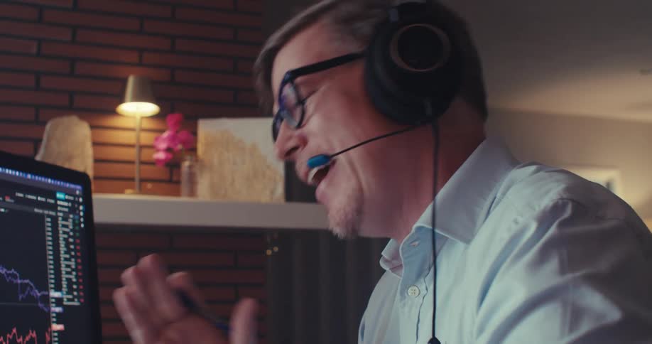 A person wearing headphones is on a call near a computer screen showing two fluctuating line graphs tracking data or financial trends