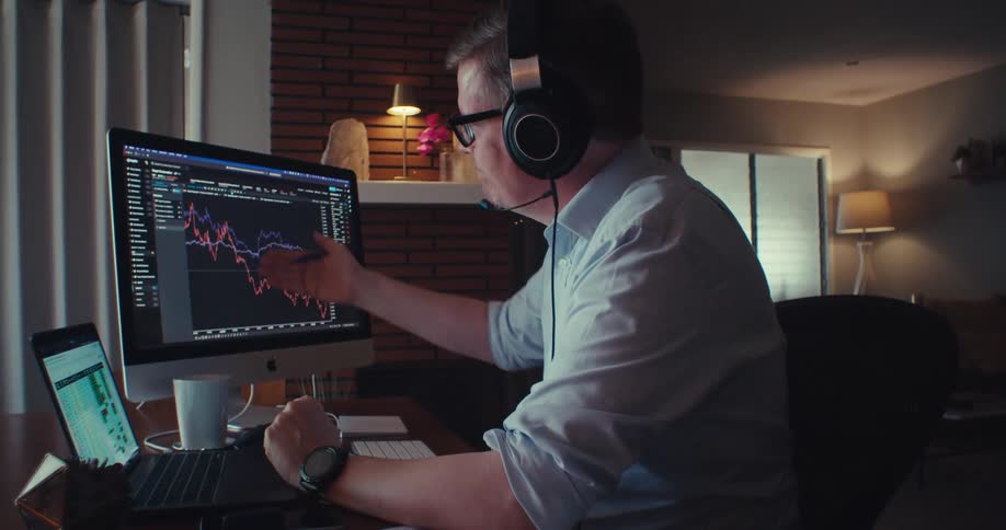 A stock trader wearing a headset sits in his living room on a conference call gesturing toward a large screen displaying live stock price charts