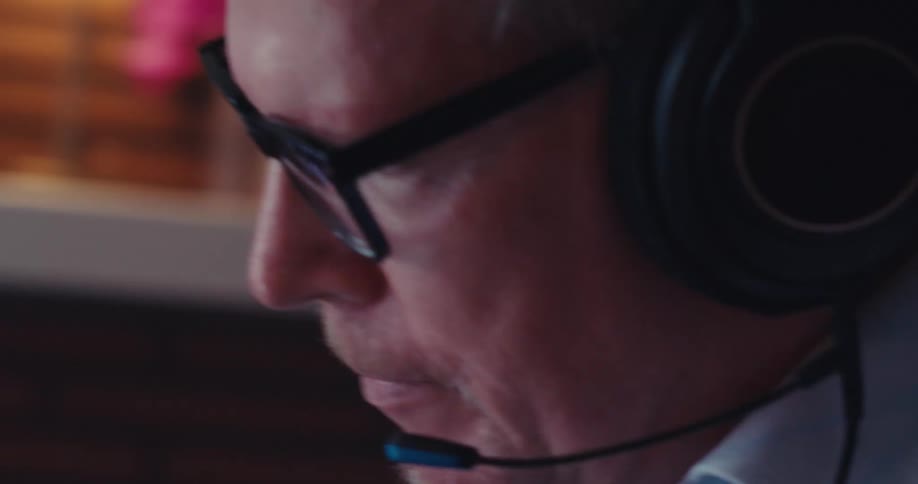 A man wearing a headset with a microphone sits in front of a computer monitor displaying financial charts speaking during a call