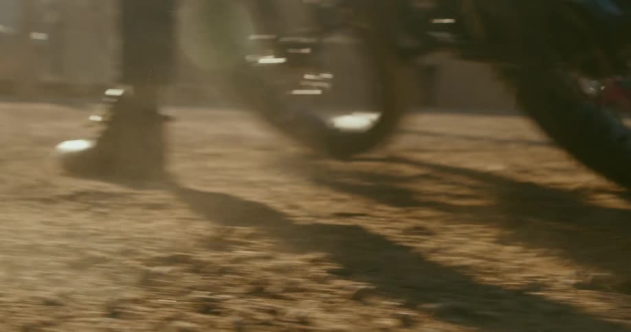 The wheels of a bike kick up dust as it spins out on gravel in a sun drenched urban environment