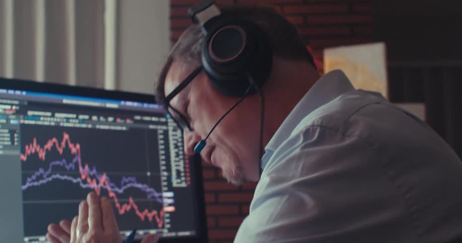 A man speaking into a headphone microphone in front of a computer screen showing stock trading in binary options