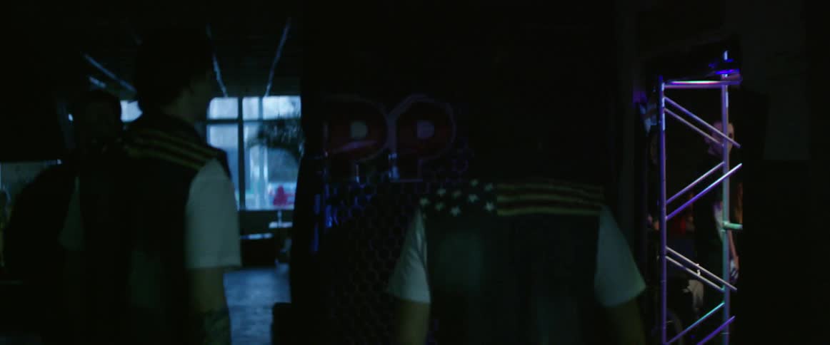 Two men in matching vests walk through a building toward a wrestling ring