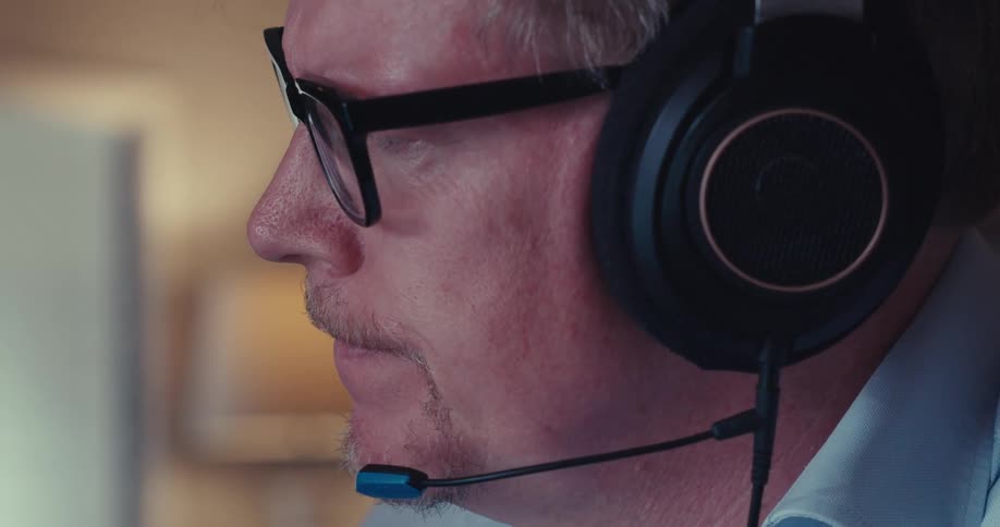 Older man wearing a headset while indoors frowns and takes off his eyeglasses