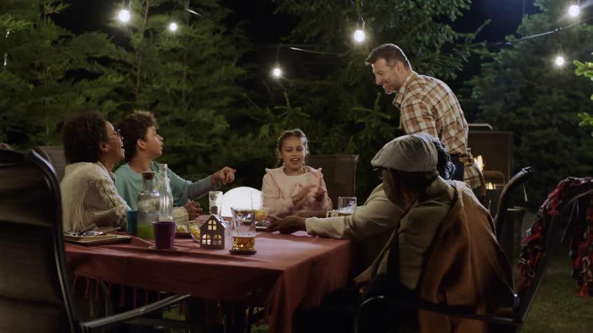 Father places grilled meat onto the table while the extended family plays a card game outdoors