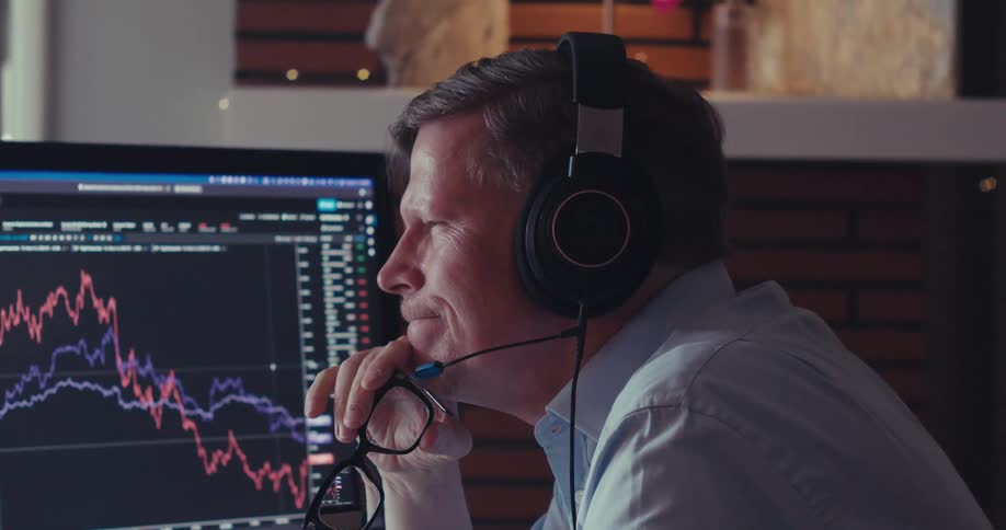A person wearing headphones sits at a desk analyzing stock market graphs on a computer screen