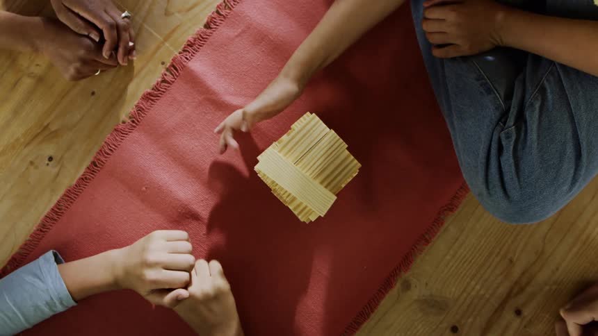Four people sit crosslegged on the floor around a red mat playing with a tower of stacked wooden blocks