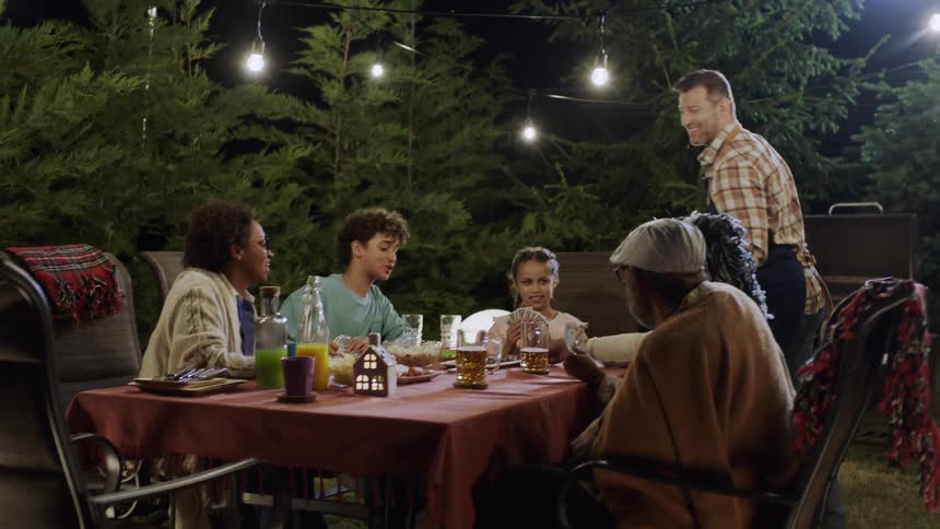 A group of people is enjoying a joyful outdoor dinner gathering under string lights at night