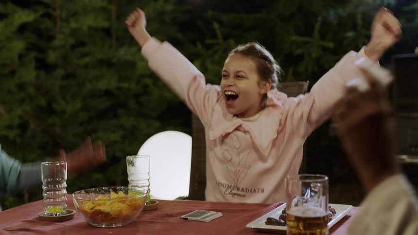 Family cheers while playing cards at an outdoor table at night