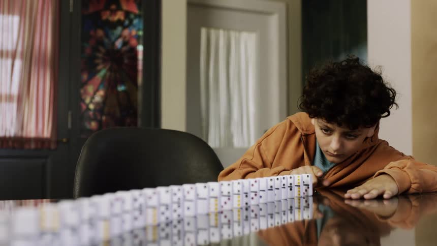 A young boy focuses intently as he sets up a line of dominoes on a table