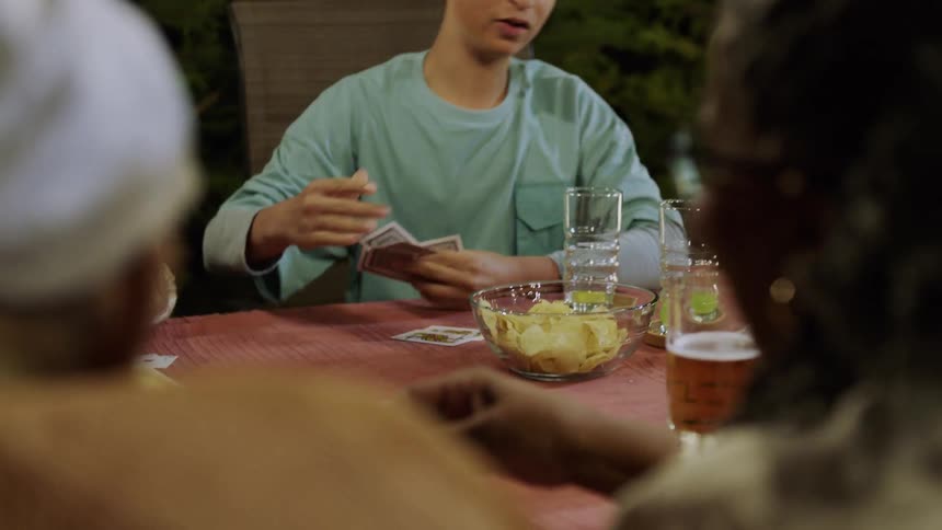 Four people play cards at an outdoor table with drinks and snacks around them