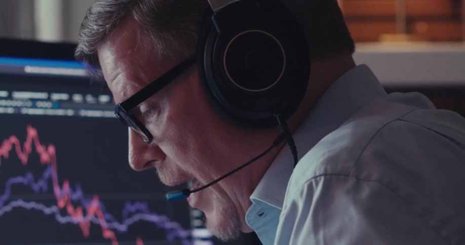 A man is speaking on a headset in front of a computer screen showing some financial information trending down