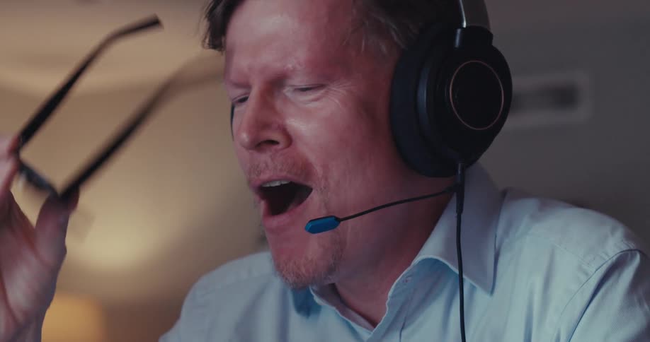 A man wearing headphones is looking down and then he starts smiling and takes off his glasses