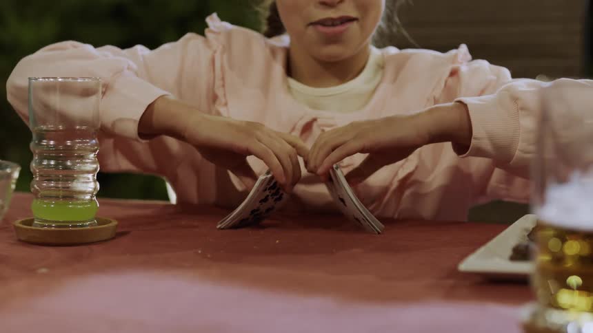 A teenage girl sits at the dining table shuffling cards laughing and having fun with a glass of drink beside her