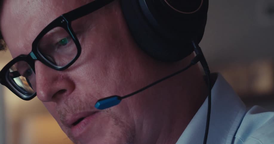 A man wearing headphones talks to someone on the other end while in his office around midday